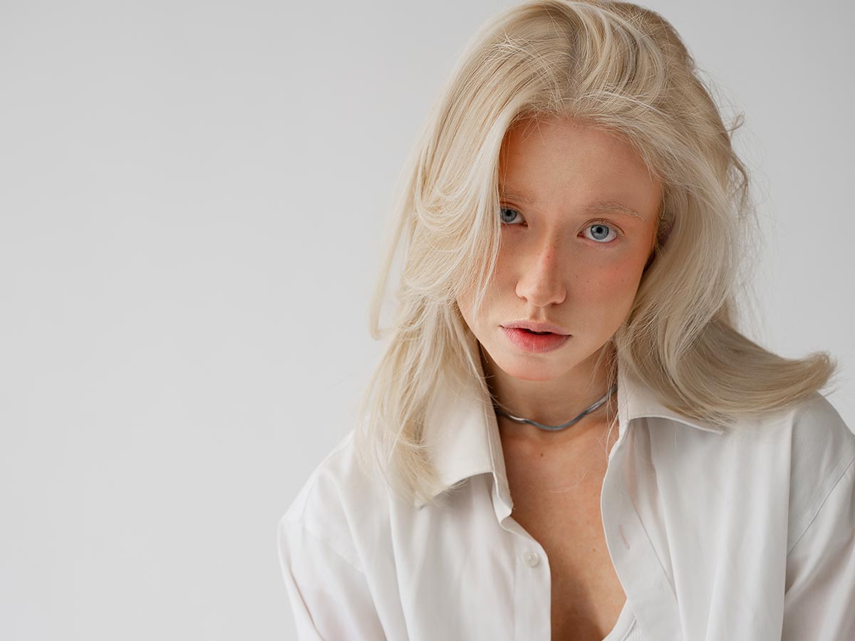 Donna con capelli biondo platino luminosi e naturali dopo l’uso dello shampoo antigiallo, indossa una camicia bianca su sfondo neutro